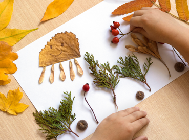 Child's hands arranging autumn leaves, berries, acorns and pine branches on white paper; one leaf is decorated with a smiley face and styled like an octopus. Scene set on a wooden table with scattered leaves.