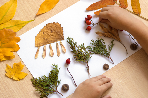 Child's hands arranging autumn leaves, berries, acorns and pine branches on white paper; one leaf is decorated with a smiley face and styled like an octopus. Scene set on a wooden table with scattered leaves.