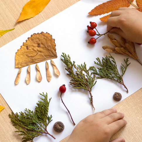 Child's hands arranging autumn leaves, berries, acorns and pine branches on white paper; one leaf is decorated with a smiley face and styled like an octopus. Scene set on a wooden table with scattered leaves.