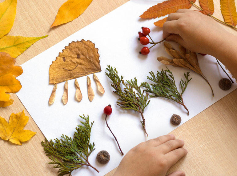 Child's hands arranging autumn leaves, berries, acorns and pine branches on white paper; one leaf is decorated with a smiley face and styled like an octopus. Scene set on a wooden table with scattered leaves.