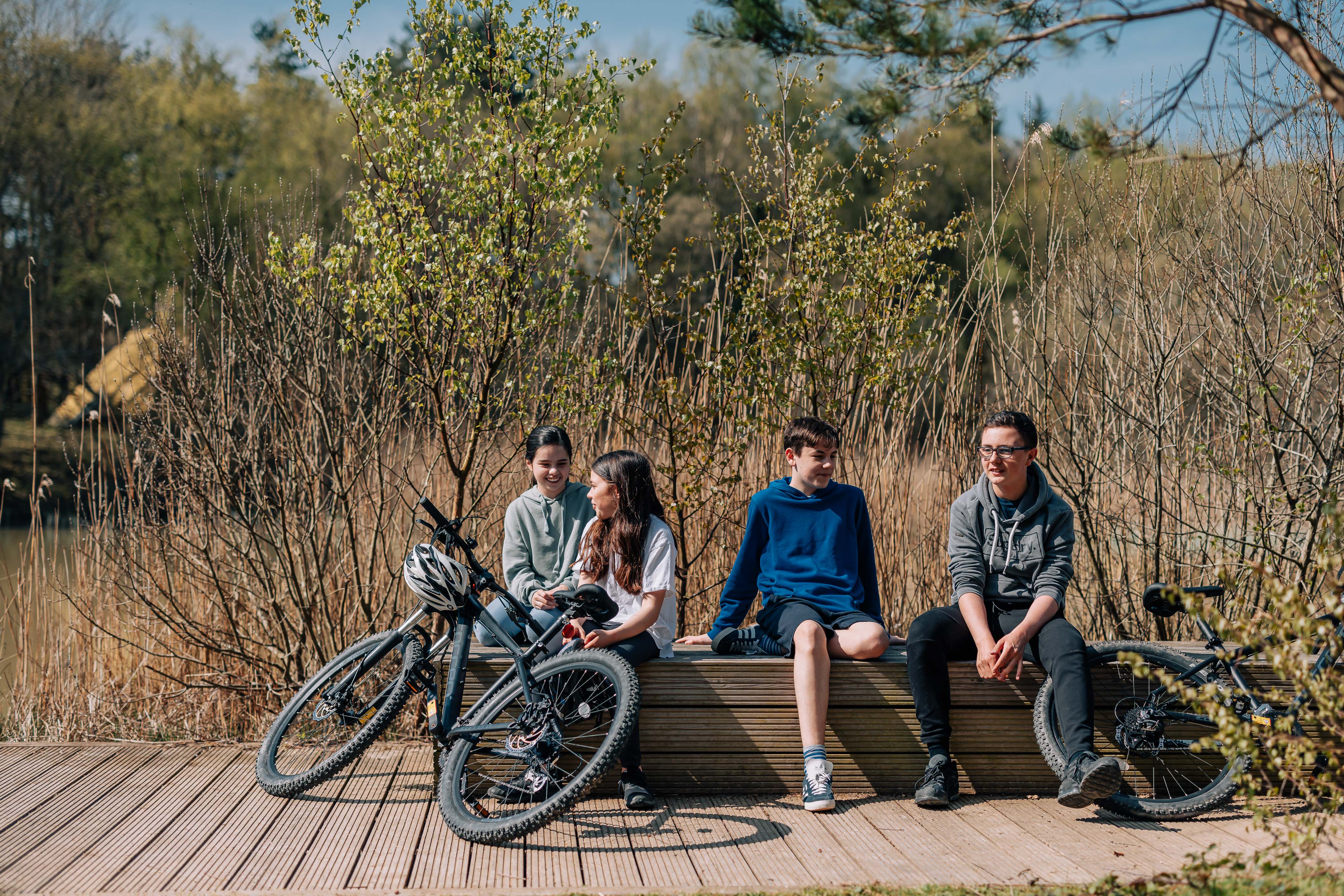 Four young people sitting on a wall with shrubs in the background and their bikes at their feet