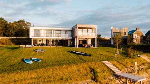 A large square modern house surrounded by grass with kayaks and paddleboards in the garden
