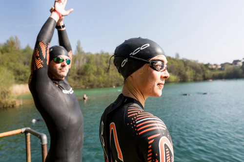 two swimmers wearing caps and wetsuits, one is stretching and getting ready to swim in a lake