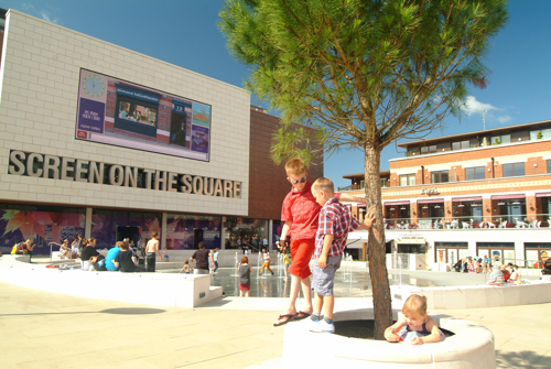 Two young boys leaning against a small tree in the town square in front of shops, restaurants and a cinema